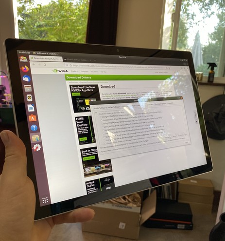 A person is holding a detached Surface Book 2 display. a webpage for downloading NVIDIA drivers. The background includes a window with greenery outside and some boxed items.