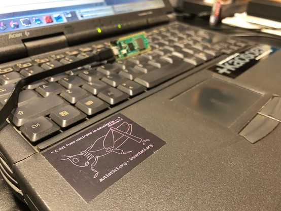 A detail of a laptop’s keyboard showing two stickers. One has a grasshopper and the writing “E dal fumo uscirono le cavallette”. The other sticker contains the “ kaos” word and both cite the autistici/inventati collective (https://www.autistici.org/)