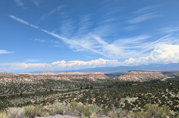 A view of two finger mesas stretching off into the distance. The rock is horizontal bands of volcanic tuff, colored in various shades of cream and red. In the distance is a mountain range with puffy cumulus clouds accumulating.