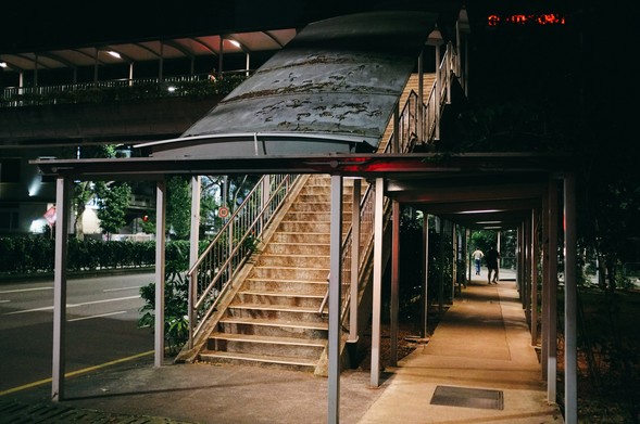 Night photo of a covered stairs leading up to the overhead pedestrian crossing on the left upper frame, and a covered walkway on the right 