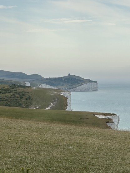 The view from the western end of the Seven Sisters over to Beachy Head in the East, with the sea on the right and green slopes on the left. The sky is a hazy blue. 