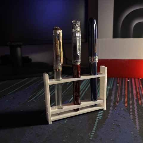 Three fountain pens within a white 3D printed stand with a dark computer monitor, a desk background, and a 3D printed flag of Poland out of focus.