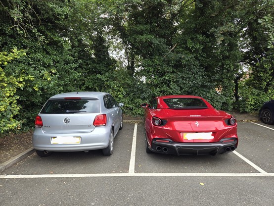 Two cars parked next to each other in a supermarket car park. The one on the left is a Volkswagen. The one on the right is a Ferrari.