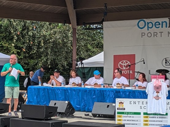 Tasting table onstage, an MC on the left and 6 people are seated. Mariatta is among the judges seated on the table.