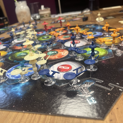 A chaotic board of hexagonal pieces with different coloured planets. Each has a counter on with a number. Between them weaves lines of model starships, occasionally broken up by a star base. The ships are coloured orange, blue, white and red.
