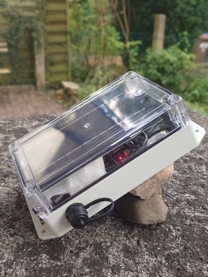 Node outside at and angle on some stones. The node and antenna are entirely in the box, and the solar panel is in the transparent top lid part.