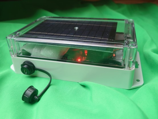 Box with transparent lid in which is a solar panel. On the side is a usb micro socket. In the box there are some red lights glowing.