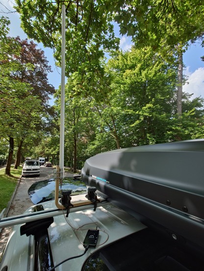 Roof of car with roof bars carrying a roof box and on the side a wooden l-shaped bracket clamped to the roof bar. The bracket carries a white antenna and the Station G2 meshastic node is on the roof of the car connected to the antenna by a thin cable. There is a USB C power cable going to the node.