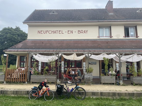 The exterior of a cafe, converted from an old station, with NEUFCHÂTEL-EN-BRAY written on the building. Several people sit on the terrace and at the front of the picture two bikes stand against the old platform edge, on what is now a grass. A young child sits on the smaller bike.