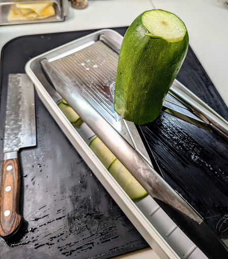 Half of the monster zucchini halfway through a slice on my large mandolin. It is placed on the bottom half of the storage case which acts as the catch container and is on a black silicon mat. The finger guard is not shown in this image but is an essential part of use. 

There is a Nakiri (Japanese vegetable slicing knife) just to the left of the mandolin which was used to trim the ends and halve the zucchini.