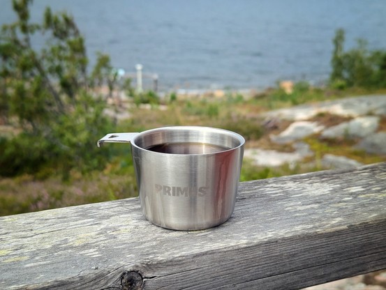 A brushed stainless steel mug, containing coffee, standing on a railing. There's some greenery and the sea in the background.