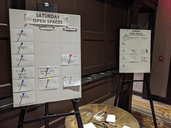 Two Poster boards: Saturday Open Spaces and Sunday open spaces, with time slots and locations for each open space. With multiple note cards in time-slots. A table with pens, thumbtacks, and papers in the bottom center.