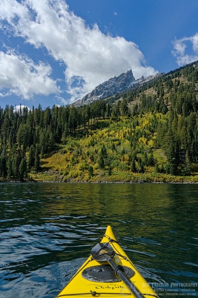 Color portrait photo of the bow of a yellow kayak on smooth greenish water pointed toward a tall sharp mountain peak in the background. In the middle ground is a conifer covered slope crossing upwards toward the right side of the frame with a patch of aspen trees and scrubs beginning to change to yellow fall colors in the middle. The blue sky is partially covered with bands of white clouds.