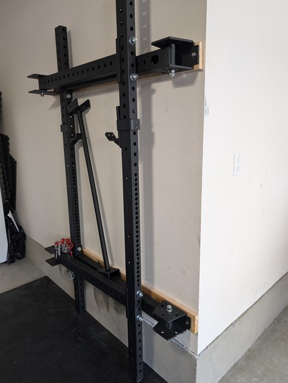 Folding squat rack, folded, against the wall of a garage, from the side.