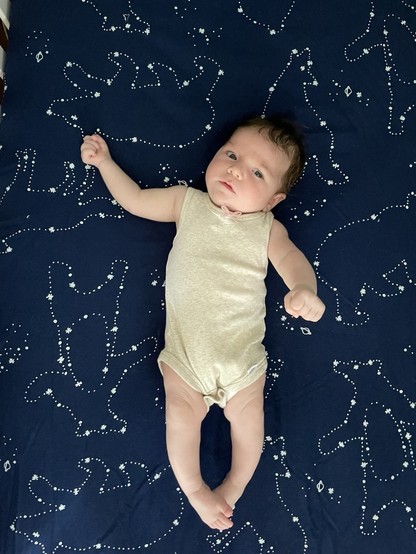 A 7-week-old baby lying on a very dark blue bed sheet. The sheet has animal constellations. The photo is taken from directly above her.
