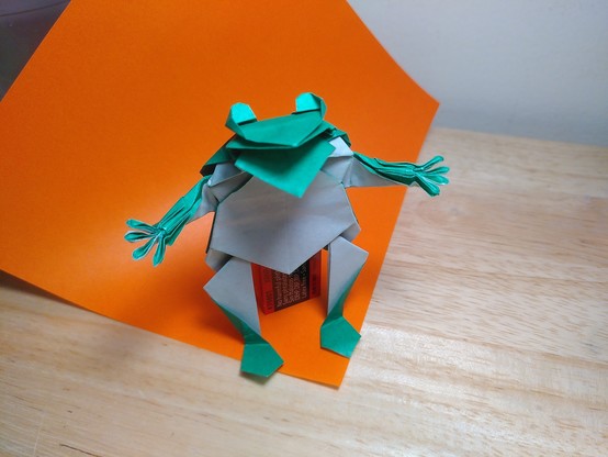 Une grenouille-bonhomme en origami qui se tient debout sur ses pattes arrières (le réalisme n'est pas ce qui est recherché dans ce modèle). Elle a quatre doigts (!) à chaque main, deux gros yeux qui lui dépassent de la tête et une grande bouche ouverte comme pour parler.