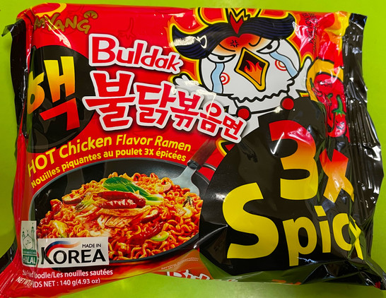 A brightly coloured packet of Buldak hot chicken flavour ramyen, 3x (3 times) spicy, sits on a violently green bench. The packet has a picture of cooked noodles in a pan, as a serving suggestion. There is a cartoon image of a crazy Korean chicken with flames in its mouth, gesturing with its wings. Net weight 140g (4.93 oz). Labelled Made In Korea, Halal.