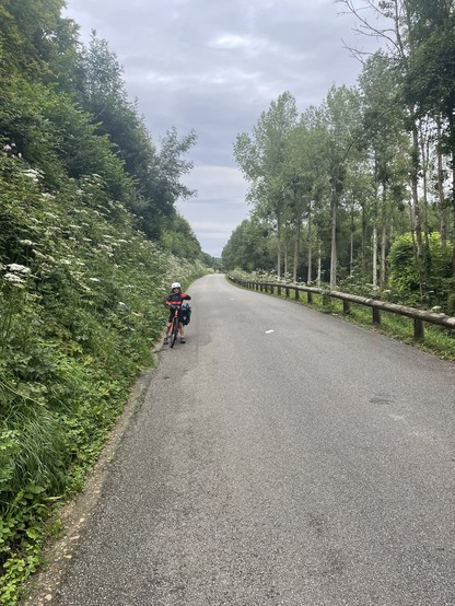 A tarmac road stretches into the distance, banked on one side by steep vegetation and a low wooden fence on the other. A young cyclists is on the edge of the road.