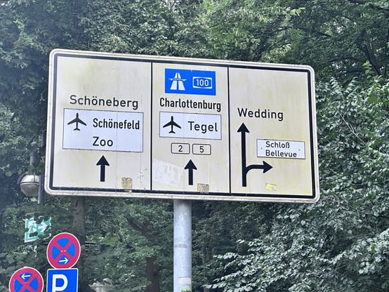 Faded yellow road sign with black arrows pointing straight ahead towards Schöneberg Schönefeld
Zoo
A-100
Charlottenburg Tegel
Wedding
And one curved arrow towards 
Schloß Bellevue in Berlin with trees crowding the background 