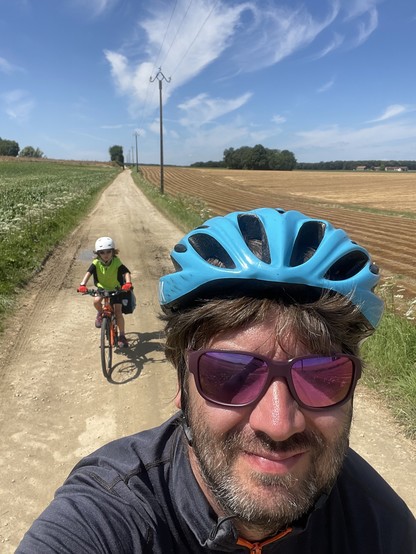 Selfie of a man in a blue bicycle helmet and sunglasses, riding on a gravel path. Behind is a child riding their own bike.