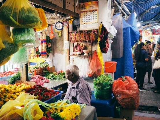 Flower garland merchant seated at his stall surrounded with different types of flowers used for garland with a puja offering shelf and a calendar on the wall behind him