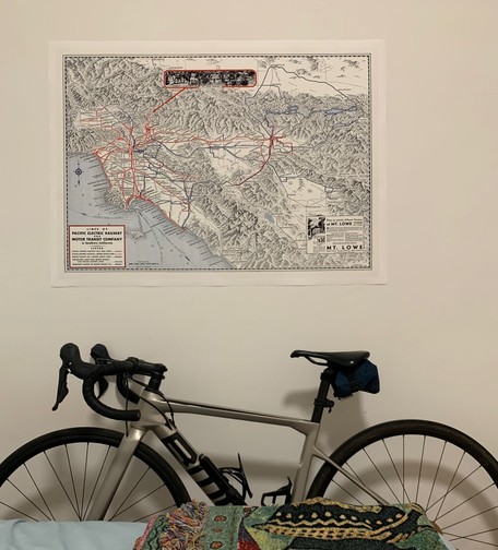 1933 Pacific Electric map showing LA-IE-OC on a white wall. A silver road bike is beneath the map leaning on the wall, partially obscured by a bed.