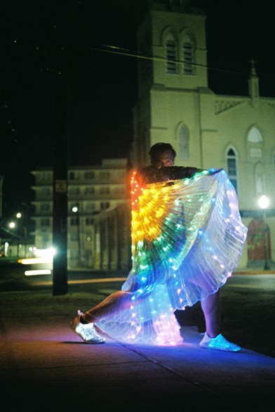 A person wearing a light-up LED rainbow cape and light-up shoes has the cape draped across their body as they strike a yoga warrior type pose at night, on a sidewalk with some buildings and street lights in the distance. 