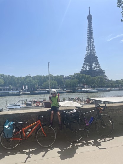 A child in cycling gear sits on top a wall beside the Seine river. Two bikes lean against the wall in front. In the background the Eiffel Tower looms.