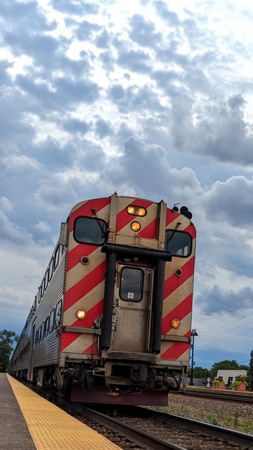 A train is approaching a platform under a gray blue sky with darker fluffy clouds. The train has two rows of windows down the side and the front has red diagonal stripes with two viewing windows with a door in the middle that has a rounded black frame giving it the appearance of a face. 