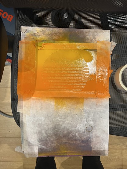 A piece of paper with orange spray paint, tape, and a stencil on it, creating a circular design with horizontal lines. The paper is placed on a black mesh chair.