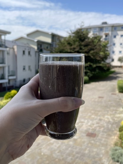A hand holds a glass of chocolate-coloured milkshake. Out of focus in the background are a wide paved path surround by shrubs and one large tree, and some three- to five-storey residential buildings.