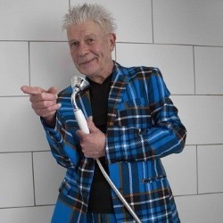 Vladimir McTavish in a tartan jacket, holding a shower handset like a microphone and pointing towards YOU