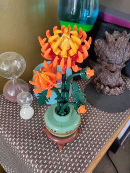 A Lego set that looks like chrysanthemums in a greeny blue vase and a wooden base. All plastic. It sits on a side table beside sand timers, a tree stump with a face and a lava lamp.