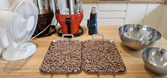 2 cooling trays each with approx 500g of roasted cocoa beans. A fan to one side blowing over them, and metal bowls to the other side ready to separate the cocoa and shells. Behind are our 2 melangers and a heat gun I was using earlier to dry the machines off as any amount of water is lethal to chocolate.