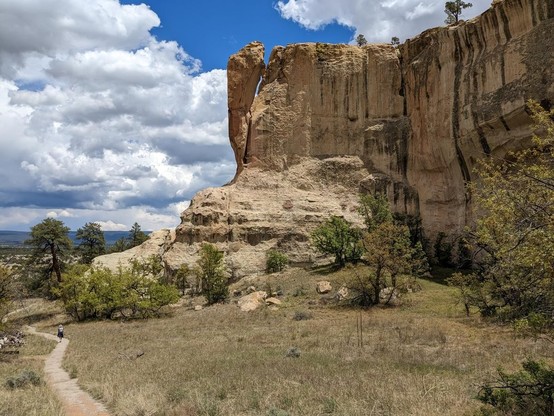 A tall cliff on the right side of the frame ends in am enormous (several hundred feet high) slab of rock that's only attached at the bottom: it looks like it could lever away from the cliff end at any moment and fall. On the left side of the frame, a trail goes past the cliff, with a tiny hiker visible on it.