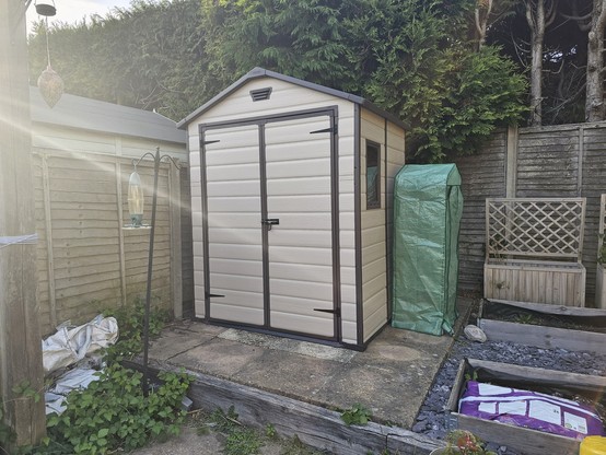 A completed composite garden shed with a mini greenhouse next to it and some flower beds