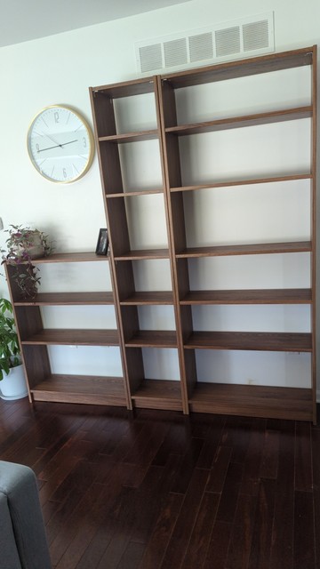 3 brown walnut color Ikea Billy bookshelves lined up against a wall with no backings so that you can see the wall behind them. Each shelf has metal corner braces screwed into the corners under the top and middle shelves to provide more structural integrity.