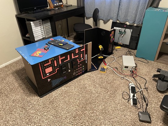 Arcade1up cabinet lying on its side, half disassembled, LCD panel in background, bench power supply and various tools strewn about. In the background are a few other pieces of “game room” paraphernalia 