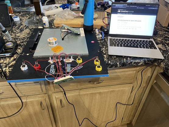 Messy kitchen table with LCD board turned upside down, soldering equipment visible, new board attached to joystick/buttons, board attached by USB to nearby laptop testing the board inputs