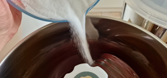 Photo of my left hand tipping a jug of weighed-out white caster sugar into the melanger