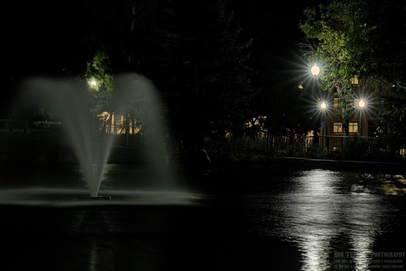 Color landscape photo of a cone shaped water fountain in a small pond on the left side of the frame. It's is night and dark. A light is backlighting the fountain. On the right side are three staggered lights on poles. They are an old fashion dome shape. Their light is also reflected in the pond. Small parts of green trees can be seen illuminated by the lights. Behind the three lights on the right are a couple of lighted windows.