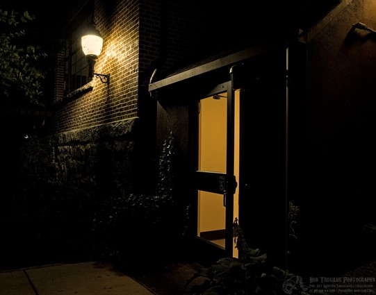 Color landscape photo of a large porch light on the left behind which is a dark window. The light is attached to a brick building with rock siding below the brick. A commercial style glass door in the center is slightly ajar. Some plants can be barely seen framing the the door. A small part of an electric conduit is seen high on the right in a bit of light cast from an unseen lamp.