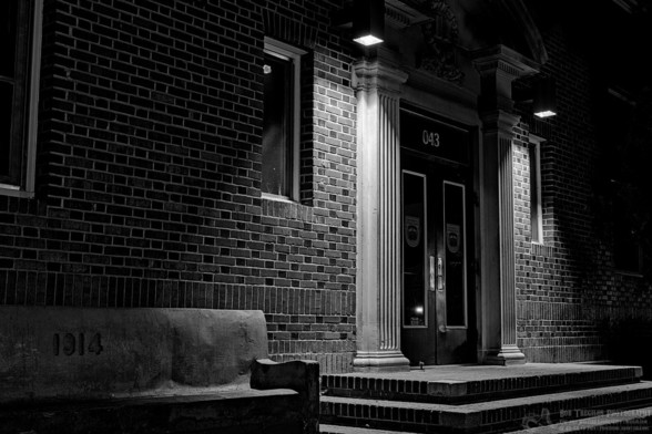 A black and white landscape photo of a former looking brick building. Twin doors are framed with faux Greek columns. Two square lights shine down from either side of the columns. The camera view is from the left at and angle. Part of a concrete bench is seen on the left in the middle of which it says, 