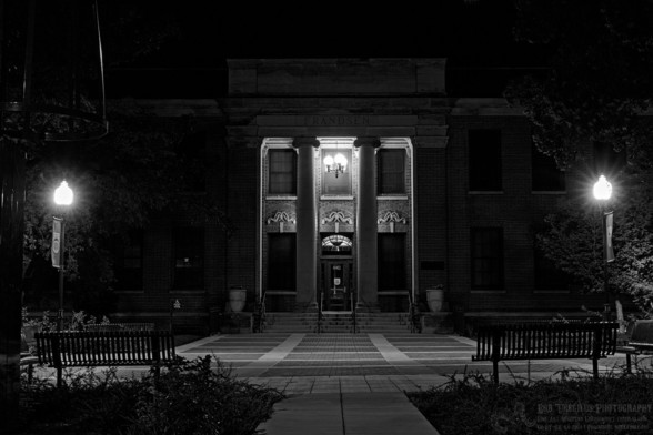 Black and white landscape photo of a formal old style building. The camera is in the center shooting towards the large front doors which are frame between two large tall columns. Steps with handrails lead up to the doors. Above the doors is a lighted eyebrow window. A set of three wing like craved decorations are on either side of the doors with one above the eyebrow window. A light with three round globes is high above the door. And there are five large windows that are dark behind the two columns. A large patio leads up to the doors with the backs of two open metal benches in the foreground. Two lights on poles are in front of the two benches.