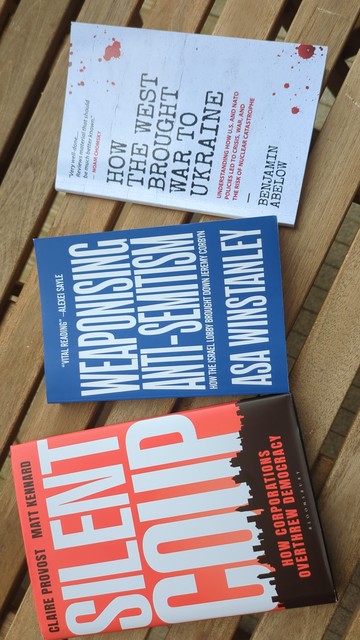 A photo of the cover of three books lay side by side on a slatted wooden garden table. Silent Coup by Claire Provost & Matt Kennard. Weaponising Anti-Semitism by ASA Winstanley. How the West bought war to the Ukraine by Benjamin Abelow.