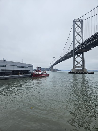 Photo of the San Francisco Bay on an overcast morning. In the foreground a red fireboat is docked at a pier in the rippled gray green water. Looming above are several spans of the Bay Bridge, stretching to Treasure Island covered with dark trees in the mid background with the east bay fuzzy on the horizon behind.