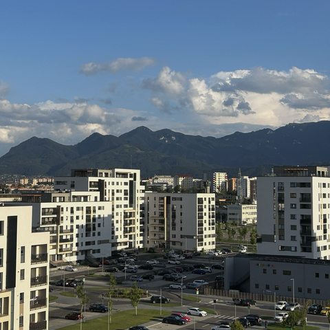 The photo features a cityscape with modern apartment buildings in the foreground. These buildings are white, multi-story structures with large windows and balconies. In front of the buildings, there are well-maintained streets and parking lots with numerous cars parked. Small patches of greenery and young trees are interspersed throughout the area, adding a touch of nature to the urban environment.  In the background, there are more buildings, gradually receding into the distance, indicating the expansive size of the city. Beyond the city, a range of tall, green mountains stretches across the horizon. These mountains have rugged peaks and are partially covered with trees and vegetation. The sky above is a clear, vibrant blue with scattered white clouds, suggesting a sunny and pleasant day.