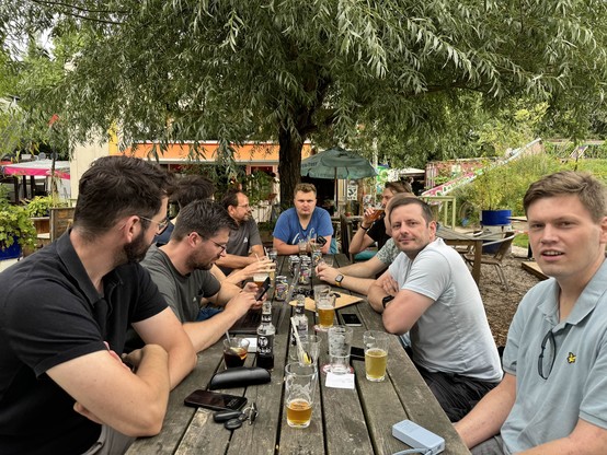 Same group of men, but they are all sitting at an outdoor wooden picnic table under a tree, from the perspective of one end of the table. A couple of the men are looking at the person taking the photo, but most are talking to each other. The table has beer glasses and bottles on it, a sign of a great time.