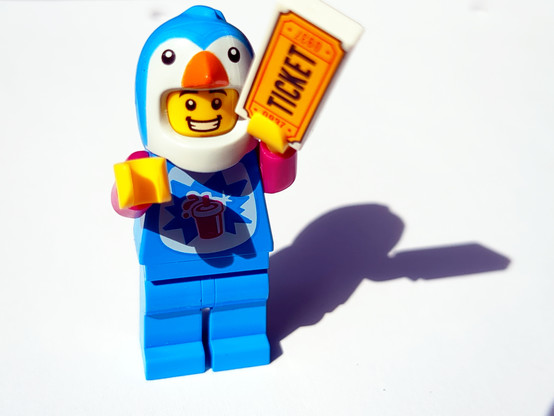 Eine Legofigur im Pinguinkostüm strahlt vor Freude und hält eine 2x1 Legokachel mit der Aufschrift 