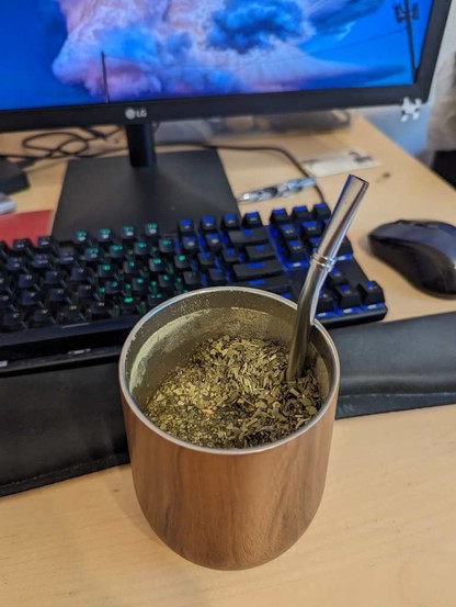 A filled stainless steel Yerba Mate gourd in front of an LED-illuminated mechanical keyboard, mouse, and monitor.